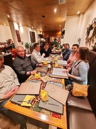 7 Personen sitzen rund um einen Holztisch mit Bast-Tischsets und gelben Servietten und braunen Menükarten. Sie sehen fröhlich und hungrig aus!